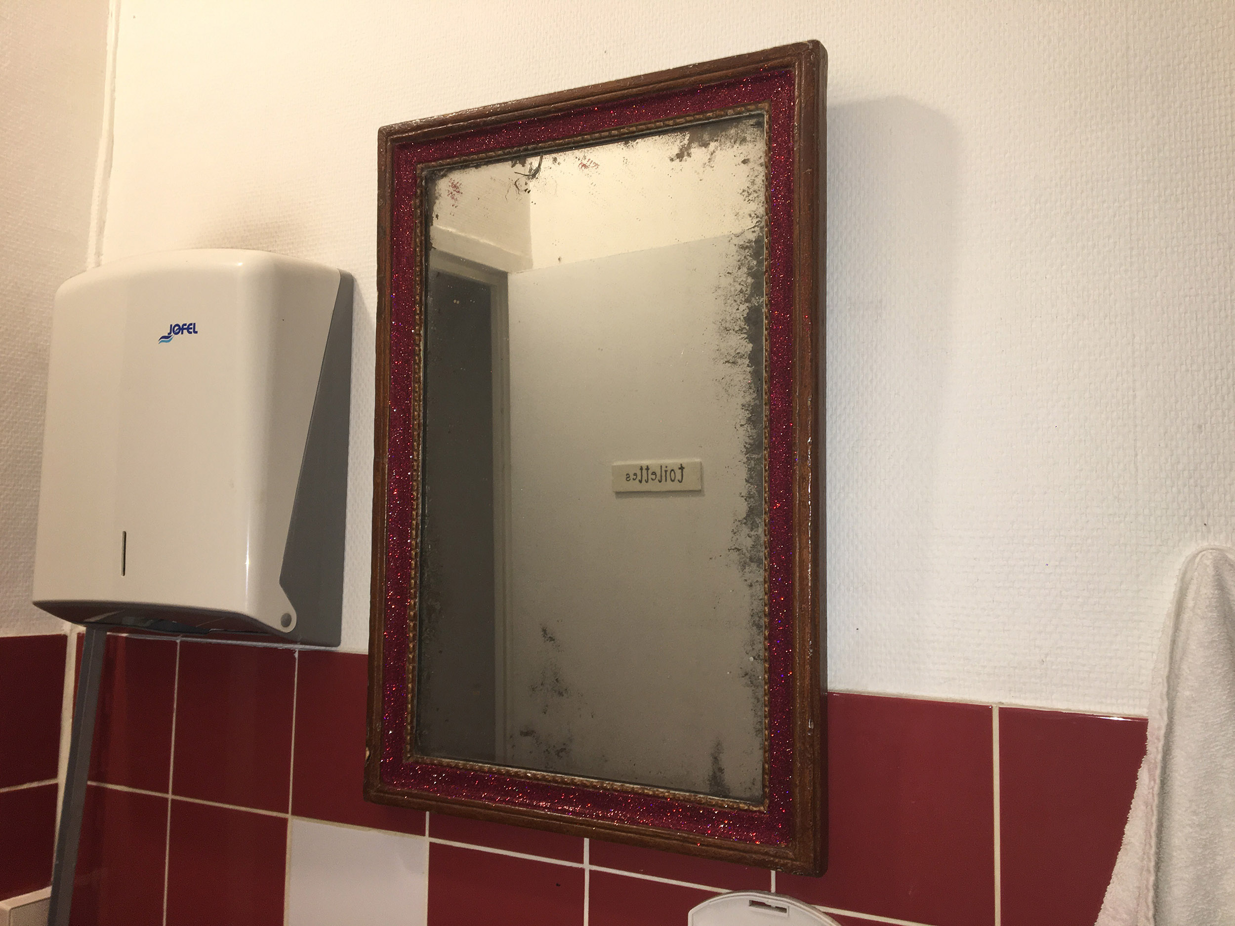 Un miroir assez ancien est installé dans des toilettes. Le cadre en bois a été recouverte de peillettes rouges.